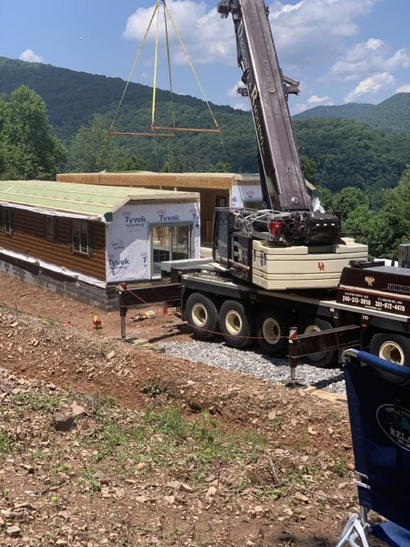 a crane setting the two pieces of a prefab log cabin together with the beautiful mountains of parson west virginia in the background