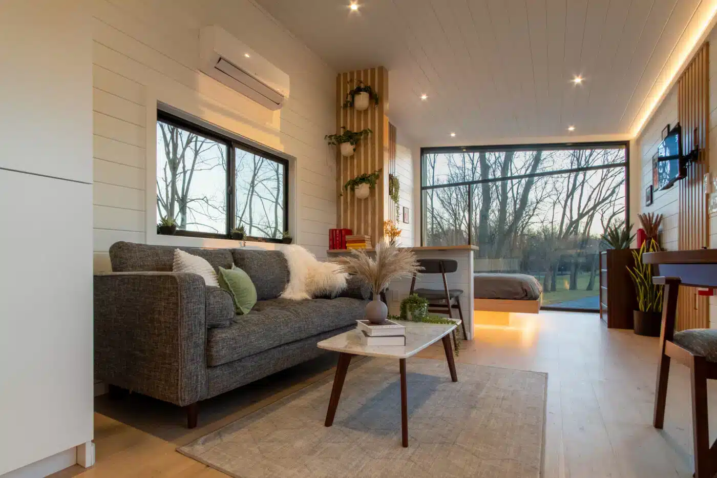 Modern tiny home interior with a cozy gray sofa, decorative plants, a small coffee table, and a bedroom area near large windows that offer scenic views of trees outside.