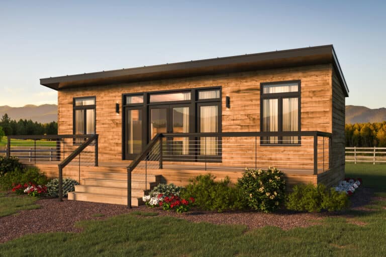 A modern Park Model Home with a sleek wooden exterior is shown. It features large windows and a spacious porch with railings, set against a backdrop of lush green grass, colorful flowers, and distant mountains under a clear sky. The home’s design is both rustic and contemporary, blending natural materials with clean lines.